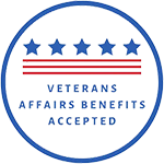 The image features a circular emblem with a blue background, containing a design resembling a shield with red and white stripes at the bottom. Inside the shield, there is text that reads VETERANS AFFAIRS SUPPORTIVE SERVICES in capital letters, followed by APPROVED in smaller font size below it. The emblem has a border with stars around its perimeter and is adorned with a red ribbon at the bottom.