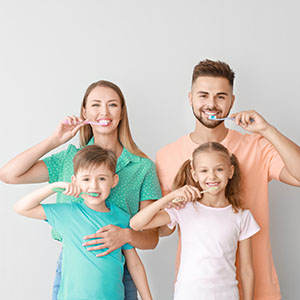 A family of four, including a man and woman with two children, brushing their teeth while smiling at the camera.
