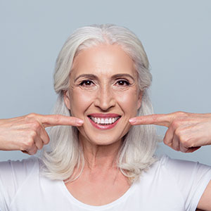 The image shows a woman with white hair smiling broadly, holding her index finger up to her mouth.